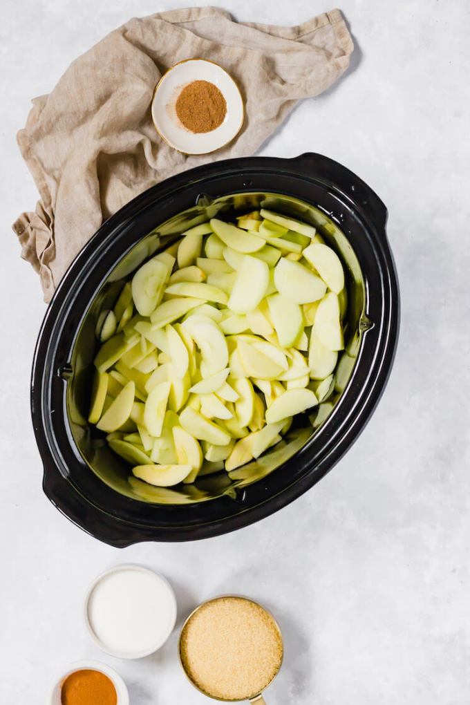 A crockpot filled with sliced apples. The rest of the ingredients for apple butter are around the crockpot.