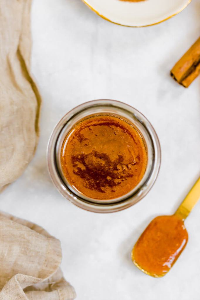 An overhead view of a jar of apple butter. A spoon with more apple butter and cinnamon sticks rest nearby.