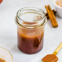 A jar filled with apple butter. A spoon with apple butter and cinnamon sticks are around the jar.