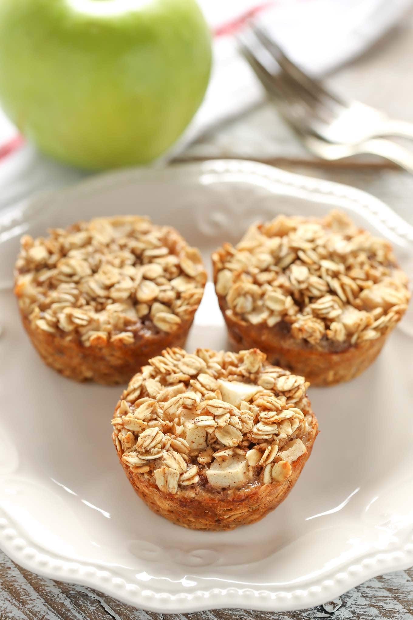 Three baked apple oatmeal cups on a white plate. A Granny Smith apple rests in the background on a tea towel.