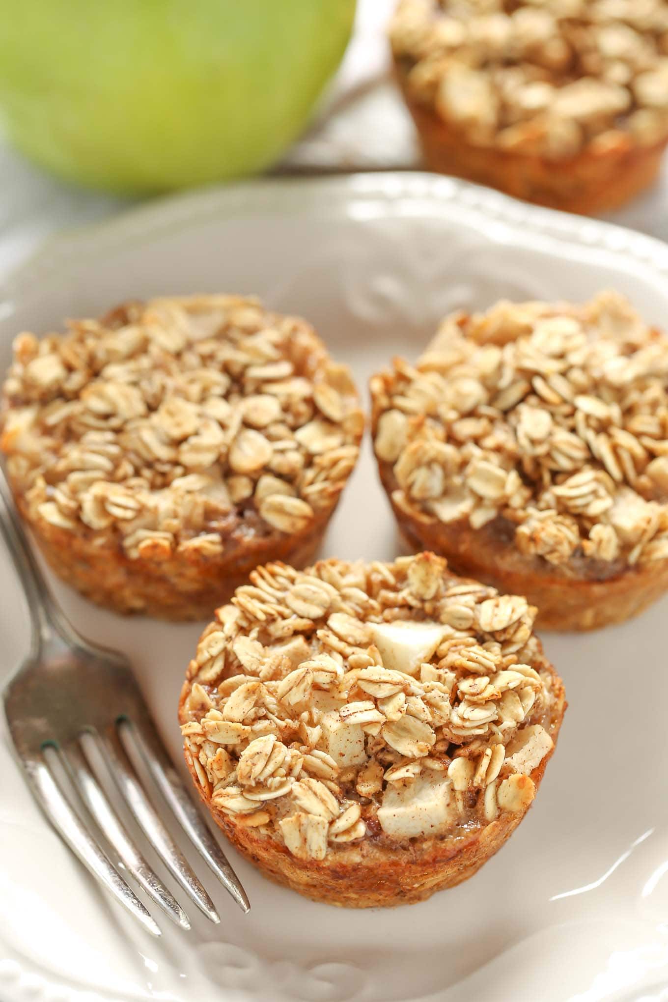 Three baked apple oatmeal cups on a white plate. A Granny Smith apple rests in the background on a tea towel.