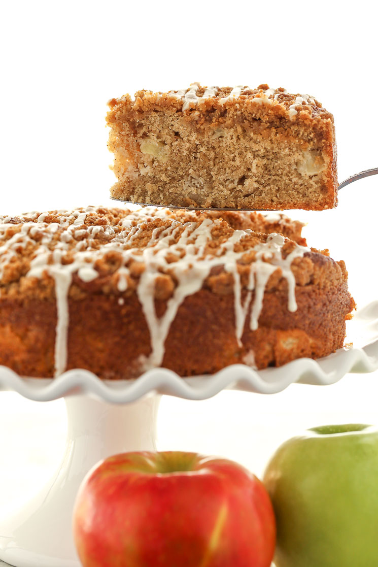 A slice of apple streusel coffee cake being lifted from a cake stand. Two apples rest in the foreground. 