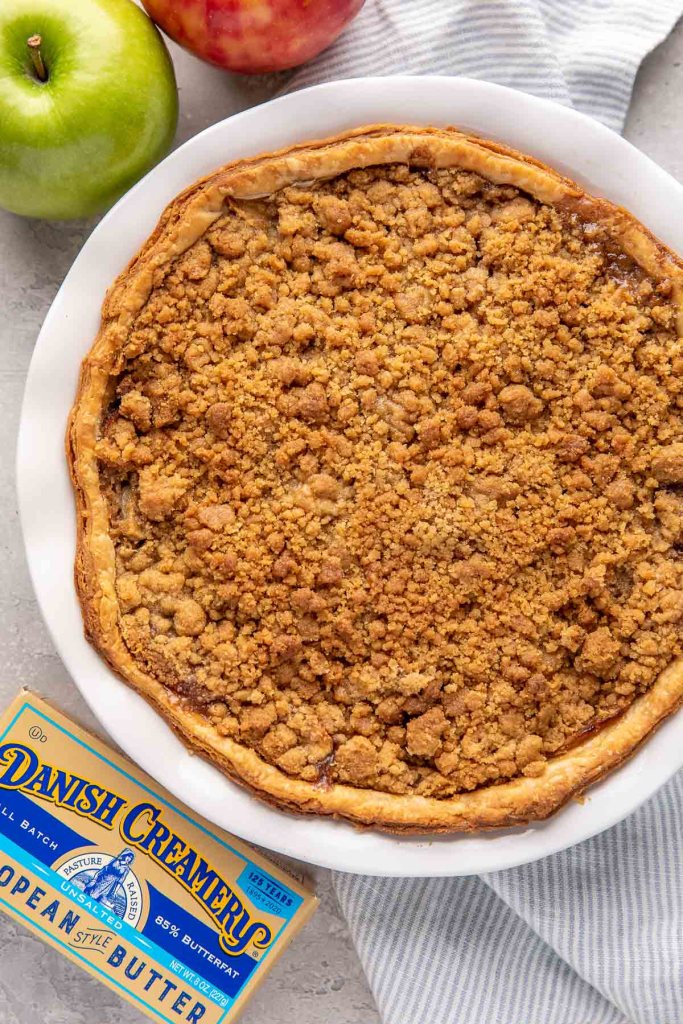 An apple crumble pie in a white pie dish. Two apples and a box of butter surround the pie dish.