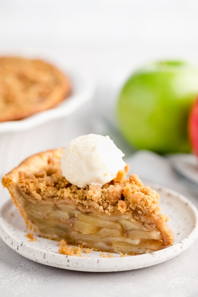 A slice of apple crumble pie topped with a scoop of ice cream on a white plate. Two apples and the rest of the pie are in the background.