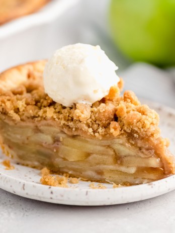 A slice of apple crumble pie topped with a scoop of ice cream on a white plate. Two apples and the rest of the pie are in the background.