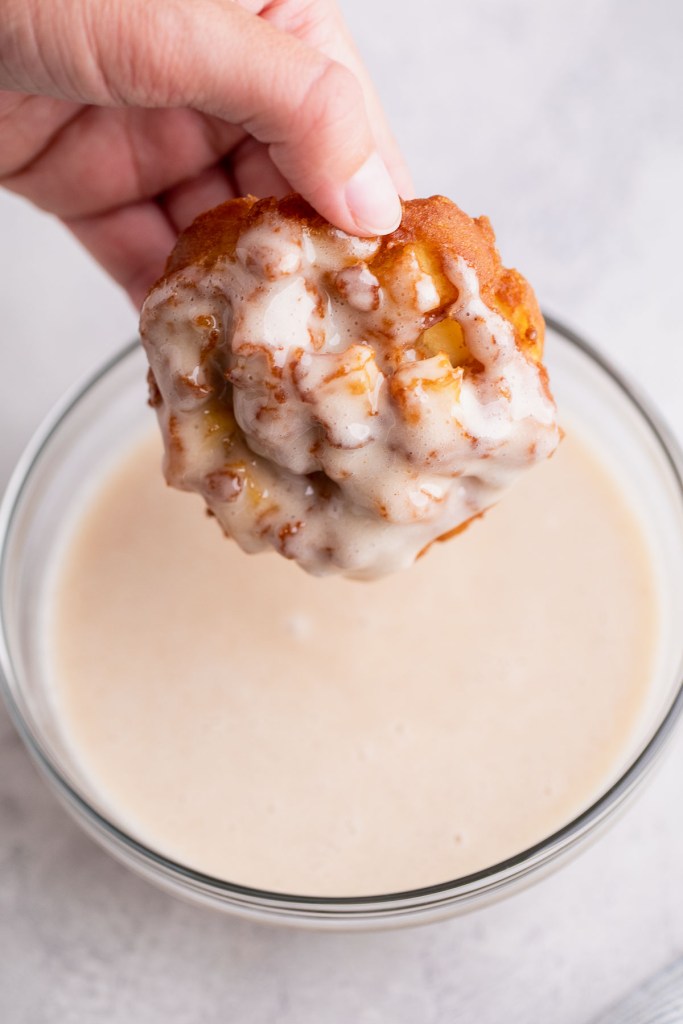 An apple fritter being held over a glass bowl of cinnamon glaze.