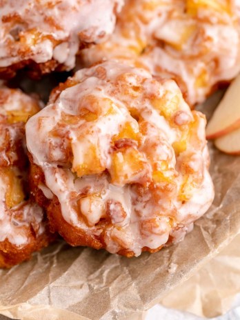 Several glazed apple fritters on top of a piece of brown parchment paper in a metal dish. A couple of apple slices are resting next to the fritters.