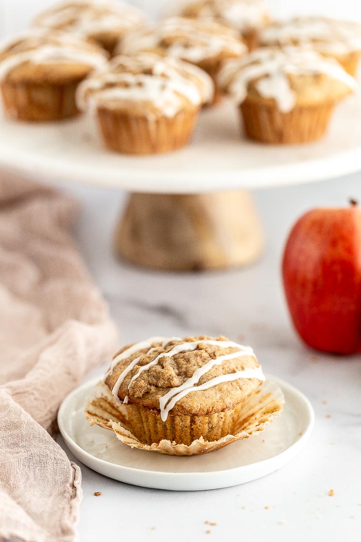 A close-up of an apple muffin on a white plate with the liner peeled away.