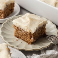 A slice of applesauce cake topped with brown butter cream cheese frosting on a scalloped plate.