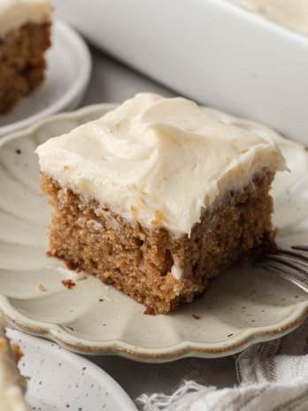 A slice of applesauce cake topped with brown butter cream cheese frosting on a scalloped plate.