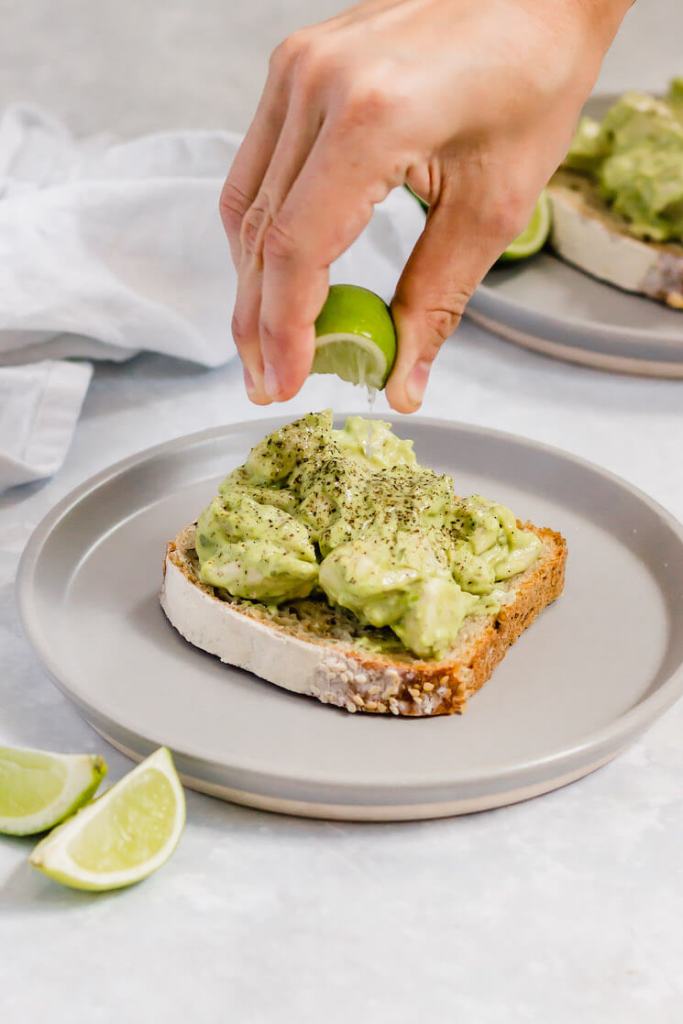 A slice of bread topped with avocado chicken salad. Fresh lime juice is being squeezed on top of it.