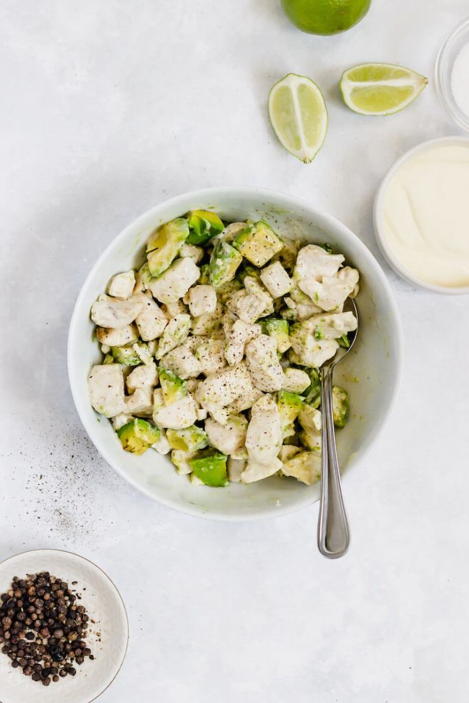 Avocado chicken salad in a white bowl. A small bowl of mayo and sliced limes are laying beside the bowl.