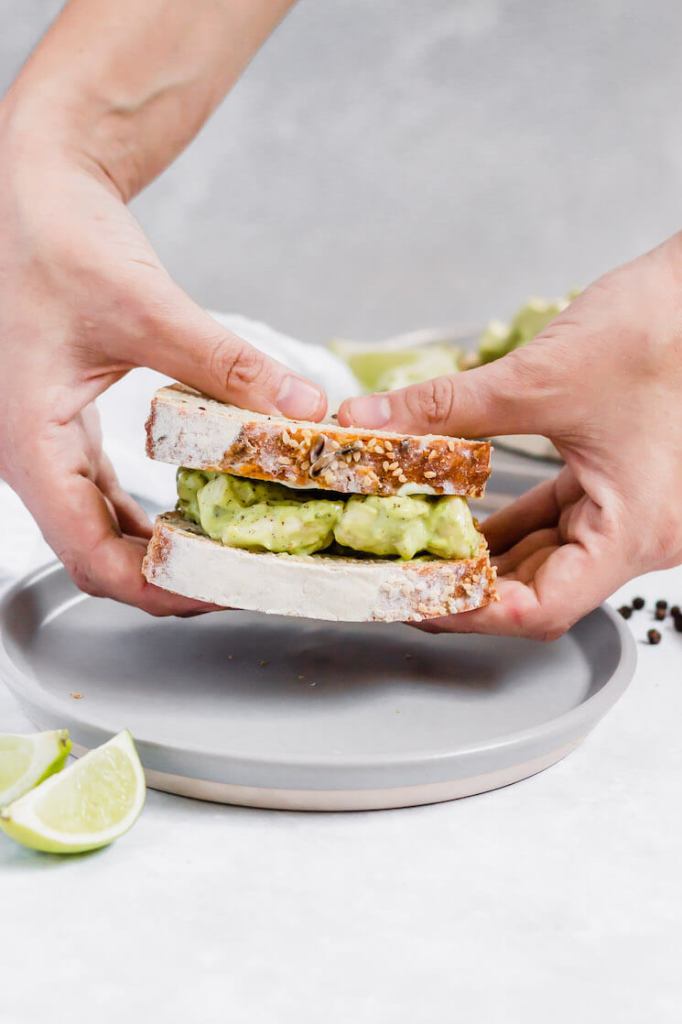 A sandwich filled with homemade avocado chicken salad. Two hands are holding the sandwich up over a gray plate.