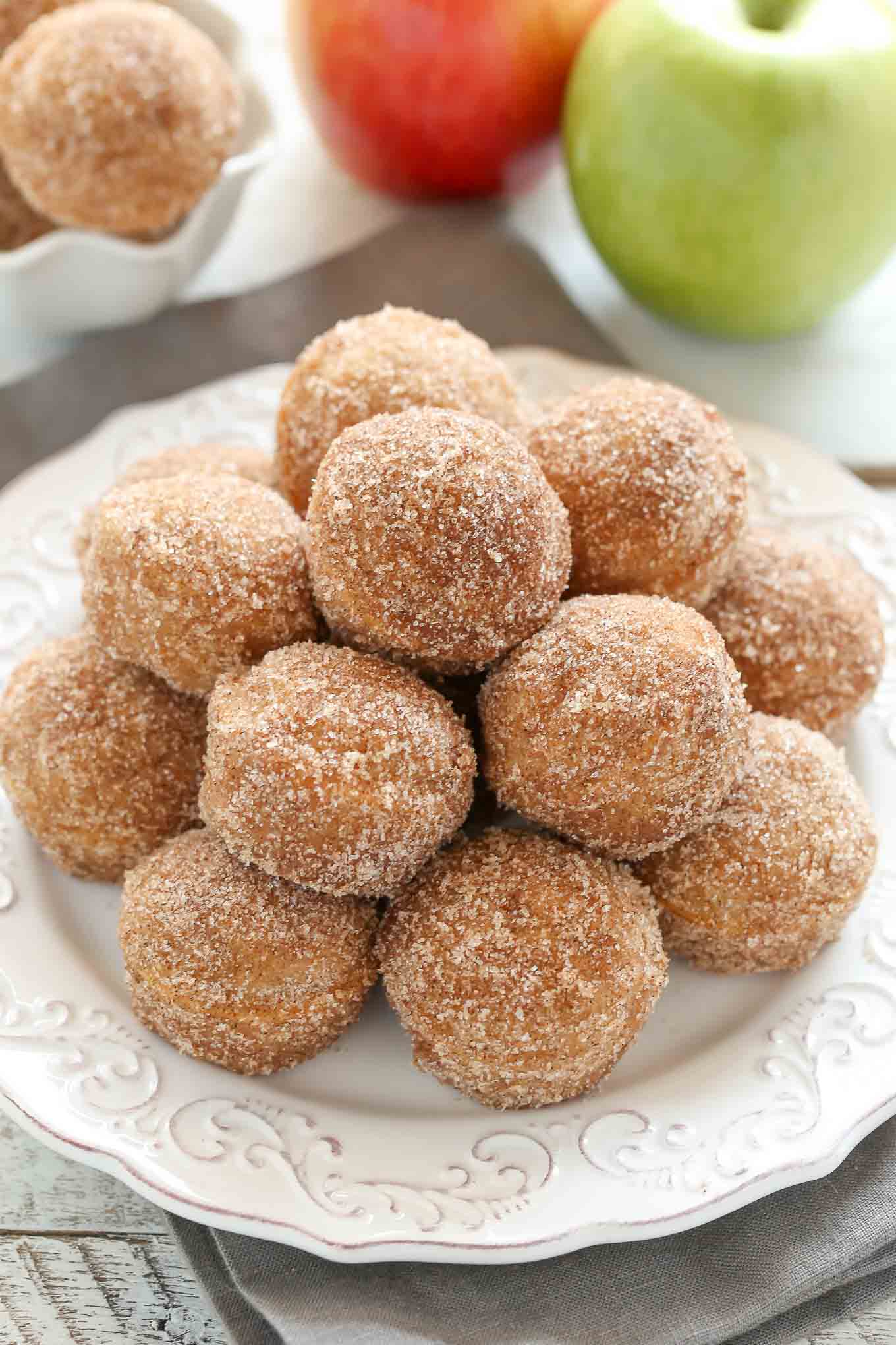 A white plate piled high with apple cider donut holes. Two apples rest in the background.