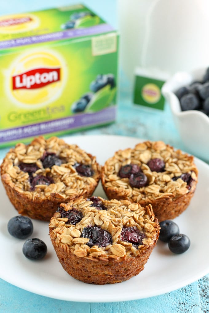 Three blueberry baked oatmeal cups on a white plate with fresh berries. A box of Lipton tea and a bowl of berries rests in the background. 