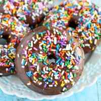 Four baked chocolate donuts topped with chocolate glaze and sprinkles on a white plate.