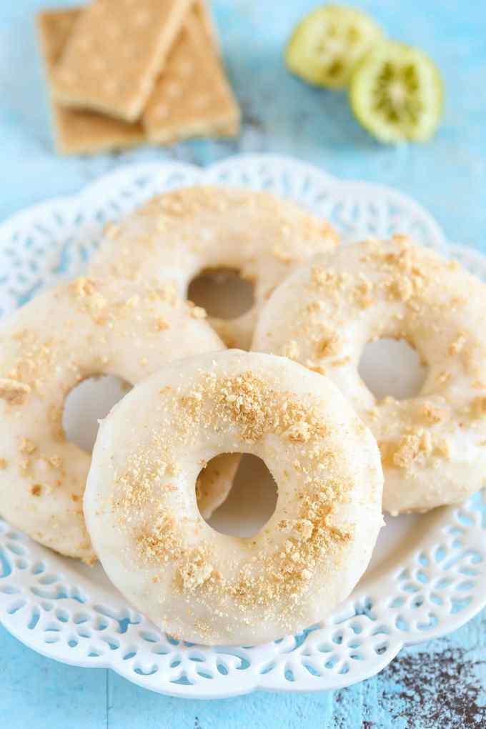 Four baked key lime donuts on a white plate. Graham crackers and lime halves rest in the background. 