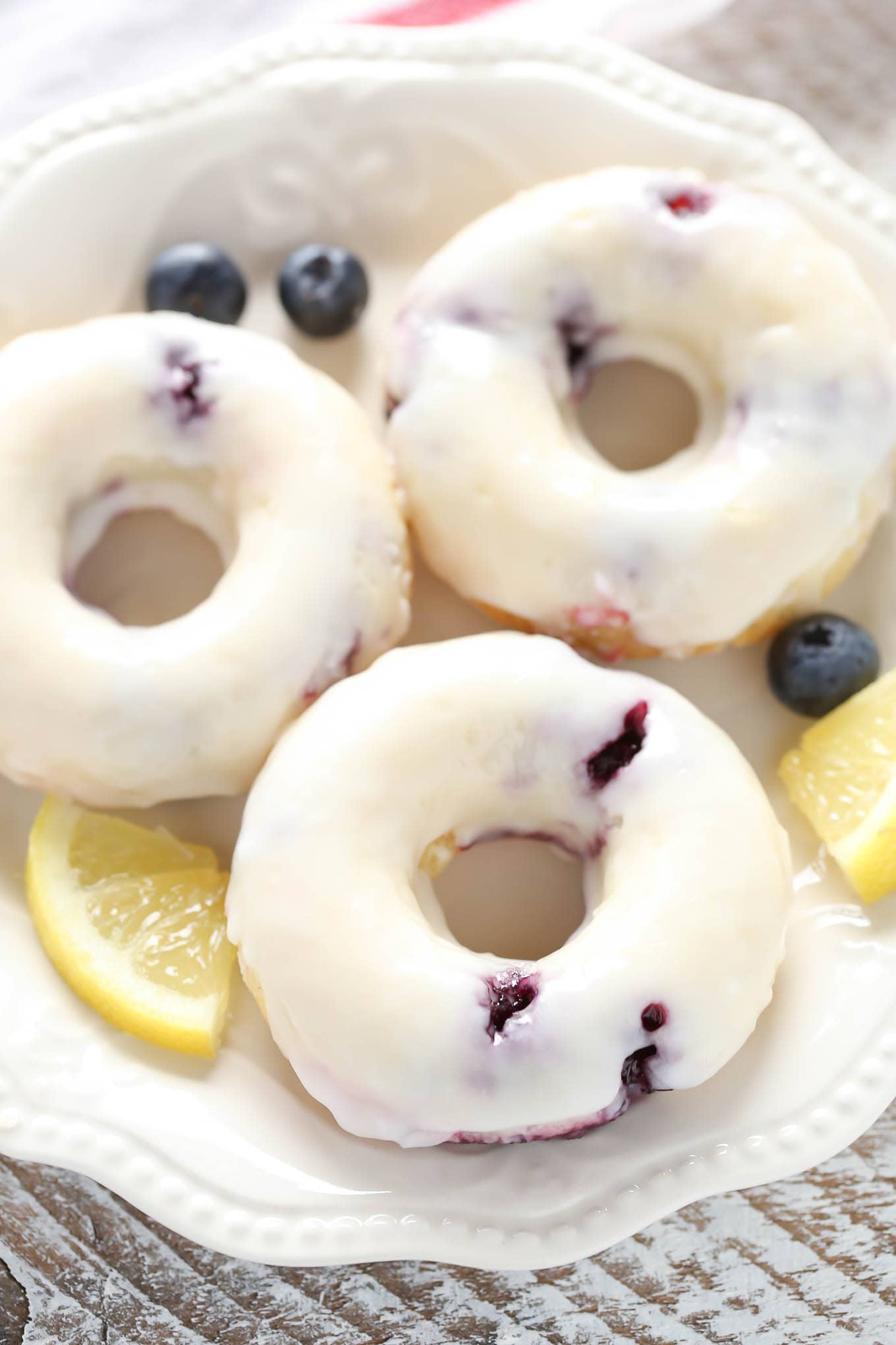 Overhead view of three lemon blueberry donuts on a white plate. Small lemon wedges and fresh blueberries surround the plate. 