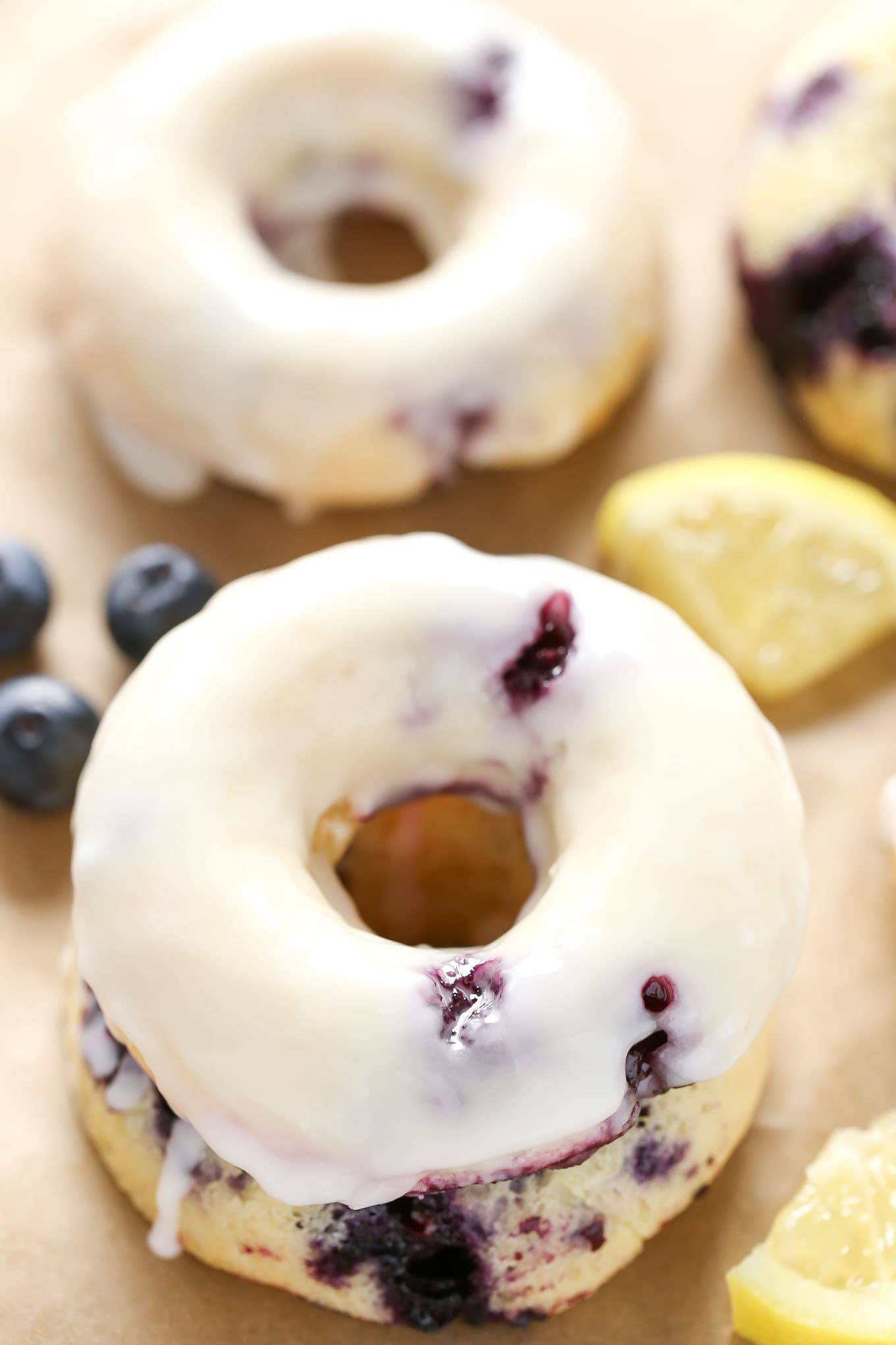 A closeup view of a lemon blueberry cake donut topped with glaze. Another donut and some berries rest in the background. 