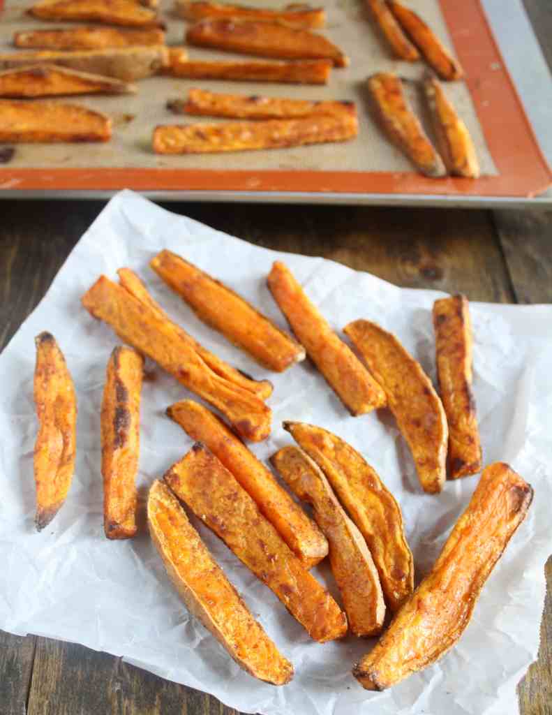 Oven baked sweet potato fries on a square of parchment paper. More fries are on a baking sheet in the background. 