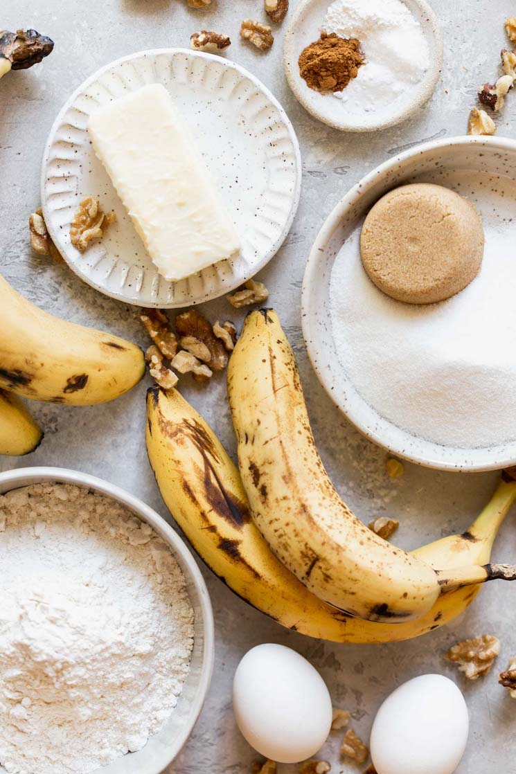 The ingredients needed to make banana bread laying on a gray surface in speckled bowls and plates.