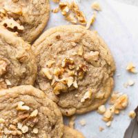 Several banana cookies on a wire rack with a piece of parchment paper underneath them. Pieces of chopped walnuts are scatter on top of and around the cookies.