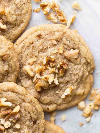 Several banana cookies on a wire rack with a piece of parchment paper underneath them. Pieces of chopped walnuts are scatter on top of and around the cookies.