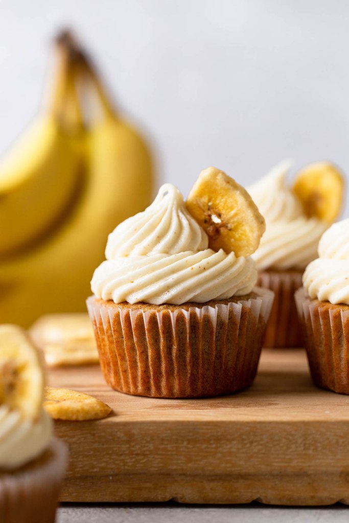 Side view of banana cupcakes with cream cheese frosting on a wooden board. A bunch of bananas rest in the background. 