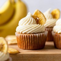 Several banana cupcakes on top of a wooden surface. Banana chips and bananas rest in the foreground and background.