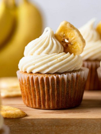 Several banana cupcakes on top of a wooden surface. Banana chips and bananas rest in the foreground and background.