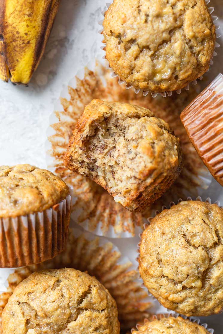 A group of banana muffins on a gray surface with one on its side and a bite taken out to show the texture. 