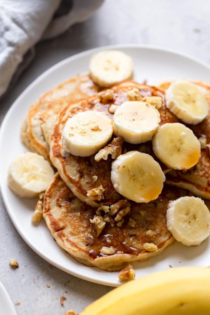 Four easy banana pancakes topped with syrup, banana slices, and walnuts on a white plate. A banana rests in the foreground. 