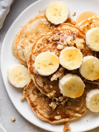 Overhead view of banana pancakes topped with banana slices and chopped walnuts on a white plate.