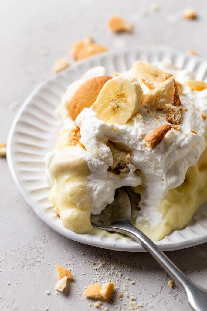 A serving of homemade banana pudding on a white dessert plate. A fork rests on the plate and crushed Nilla Wafers surround it. 
