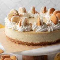 A banana pudding cheesecake on top of a marble cake stand. The cheesecake is topped with swirls of whipped cream, sliced bananas, and Nilla wafers.