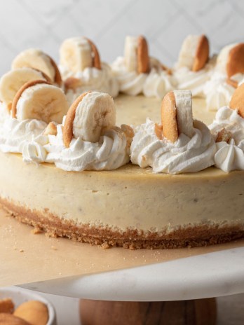 A banana pudding cheesecake on top of a marble cake stand. The cheesecake is topped with swirls of whipped cream, sliced bananas, and Nilla wafers.