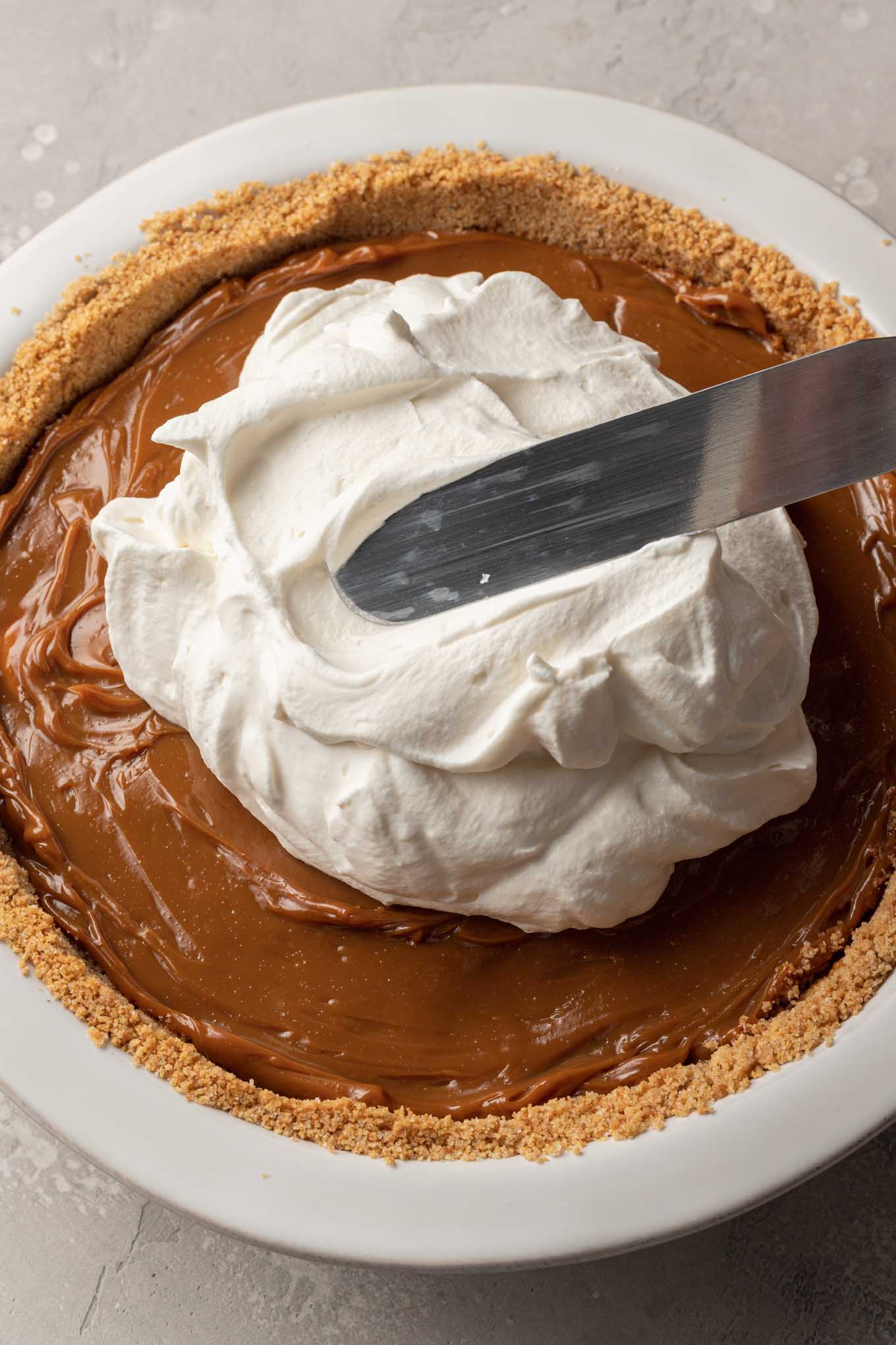 Whipped cream being spread over dulce de leche in a pie dish.