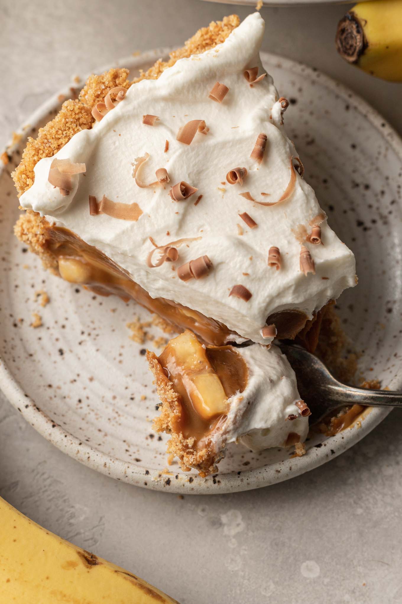 A slice of banoffee pie on a speckled dessert plate, with a bite speared on a fork. 