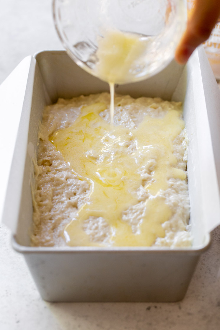 A metal loaf pan holding the beer bread batter with extra butter being poured on top.