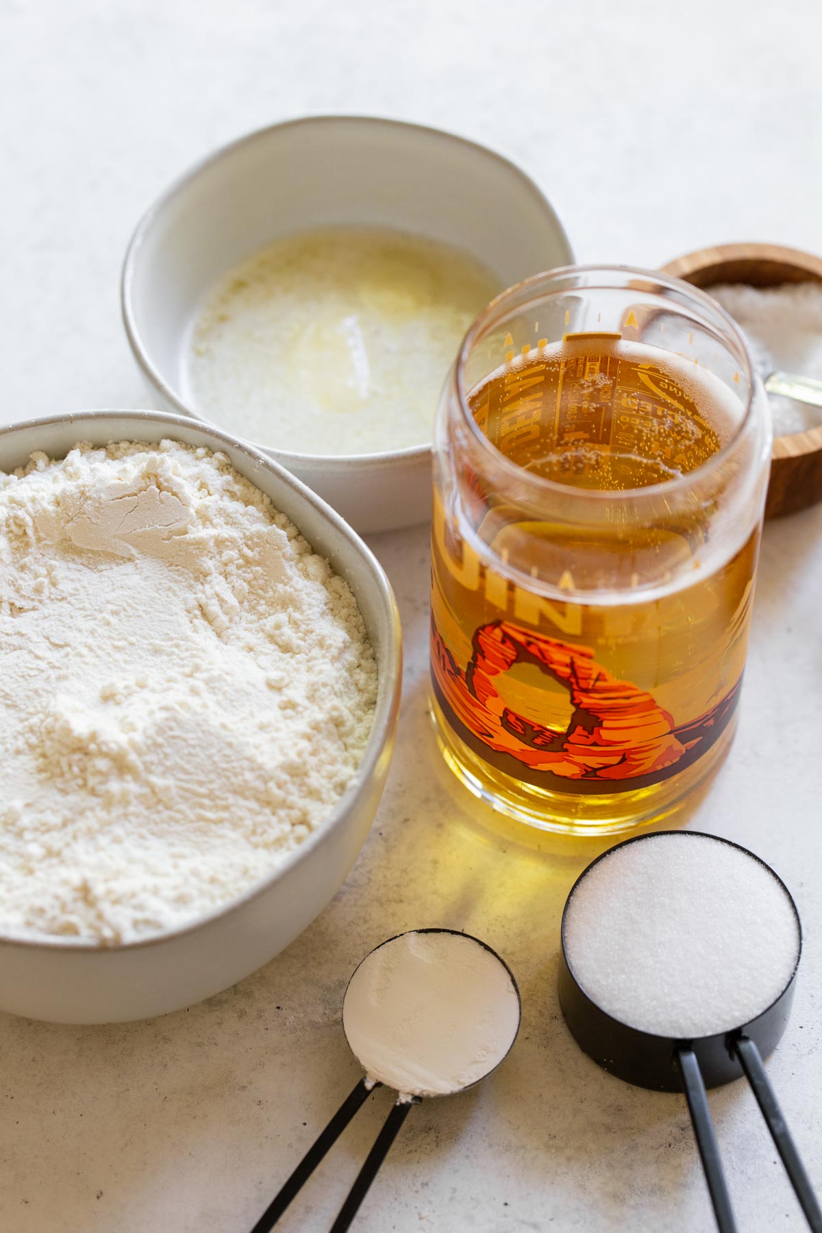 The ingredients needed to make beer bread displayed on a rustic gray surface in a variety of bowls and cups.