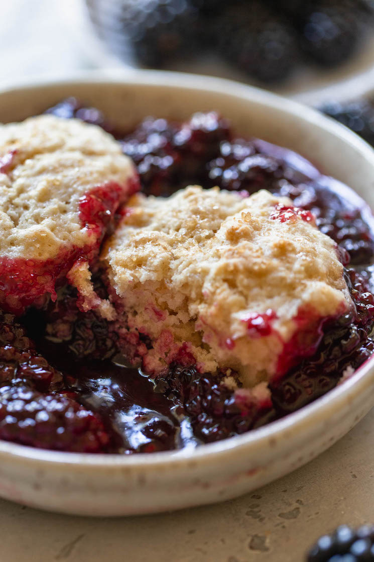 An antique bowl holding blackberry cobbler with a bite taken out of the biscuit topping to show the texture.