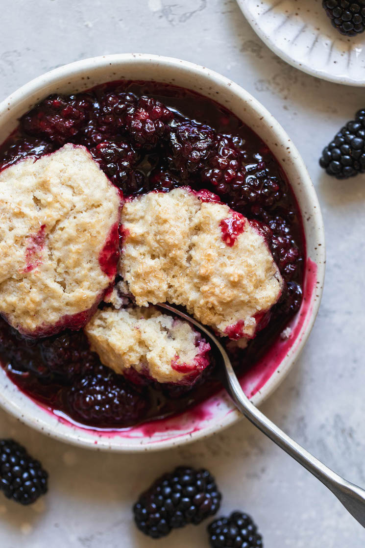 An antique bowl filled with blackberry cobbler and a spoon taking a bite out.