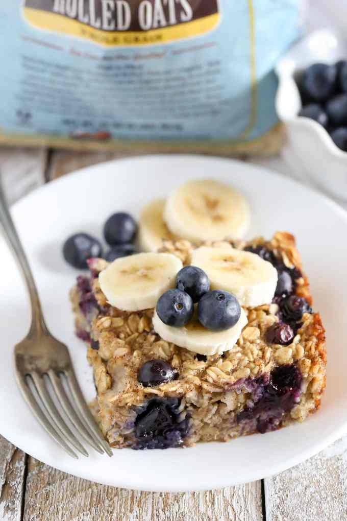 A slice of banana blueberry baked oatmeal garnished with banana slices and fresh blueberries on a white plate with a fork.