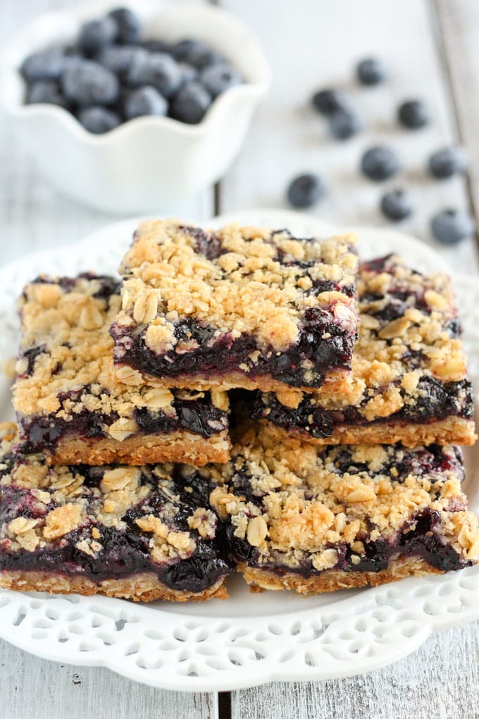 A stack of blueberry crumble bars on a white plate with a dish of fresh blueberries in the background. 