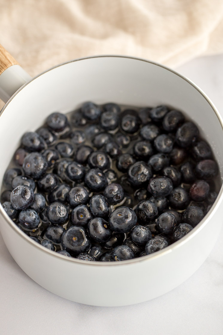 A white saucepan filled with fresh blueberries, water, sugar, and lemon juice.