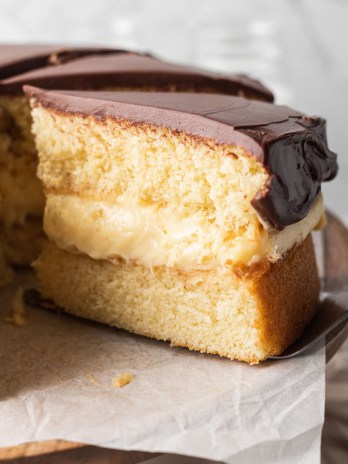 A sliced Boston cream pie on a cake stand. One slice is being removed.