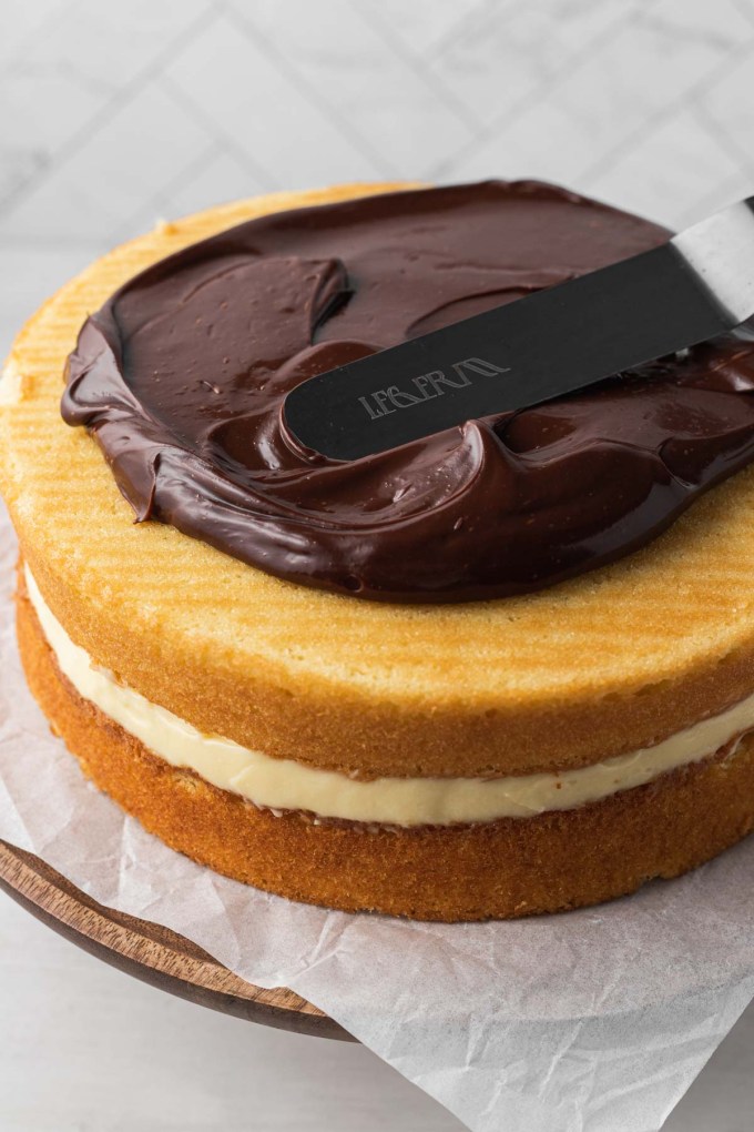 Chocolate ganache being spread over a homemade Boston cream pie cake.