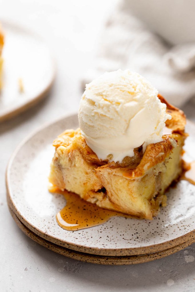A slice of bread pudding topped with a scoop of ice cream on a white speckled plate.