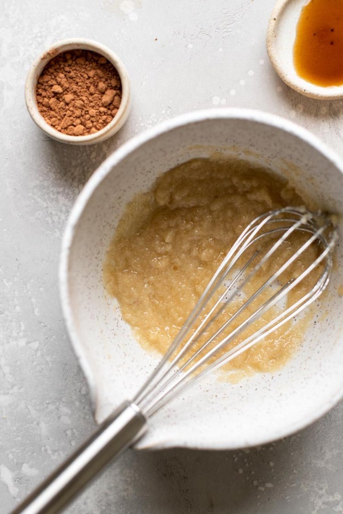 Melted butter and sugar in a mixing bowl with a whisk. A small dish of cocoa powder and vanilla extract sit on the side.