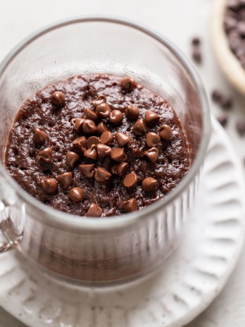 Overhead view of a microwave mug brownie in a glass mug. The mug sits on a white dessert plate, and a dish of mini chocolate chips rests on the side.